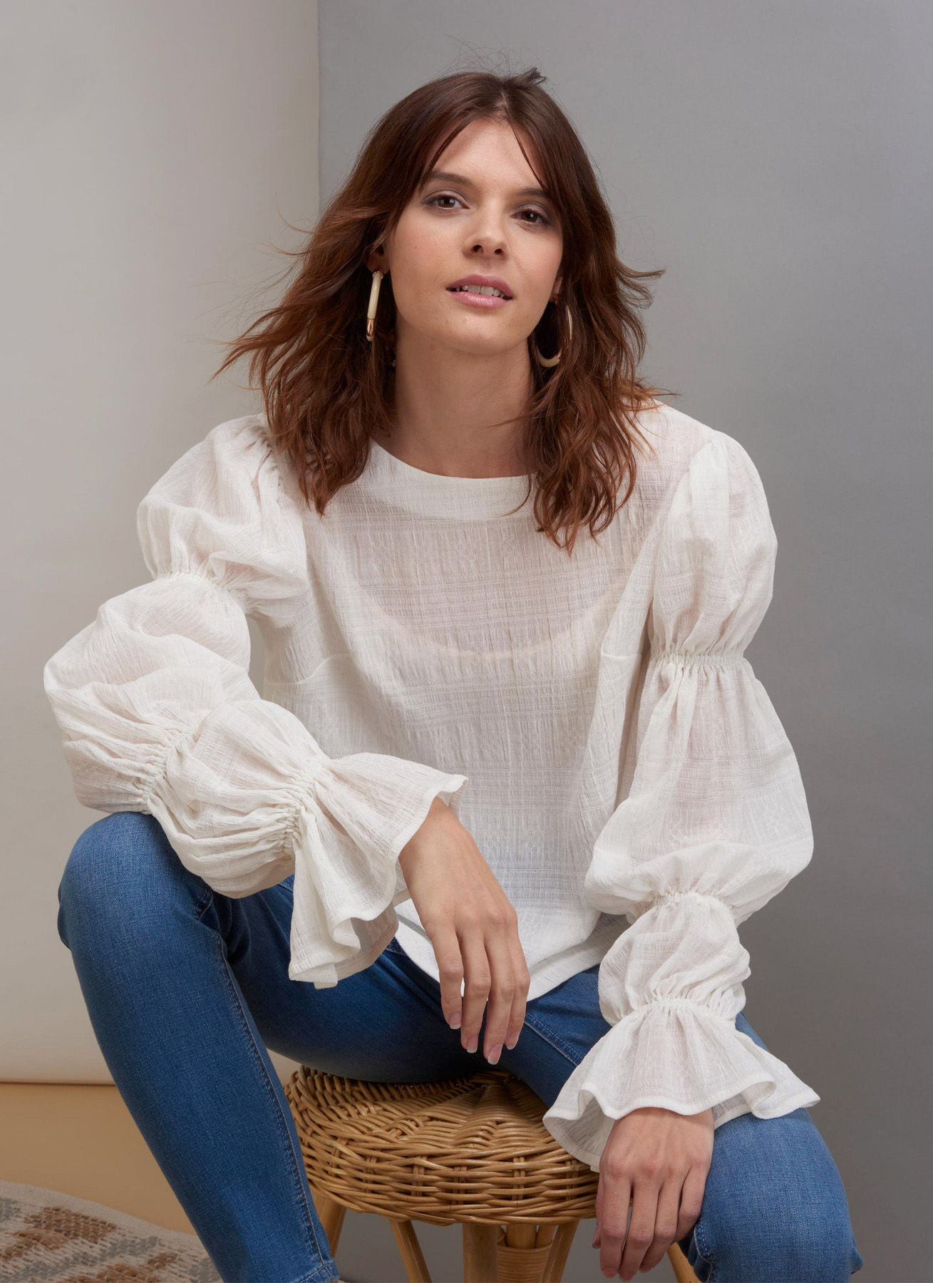 Woman in a white blouse and blue jeans sitting on a woven chair.