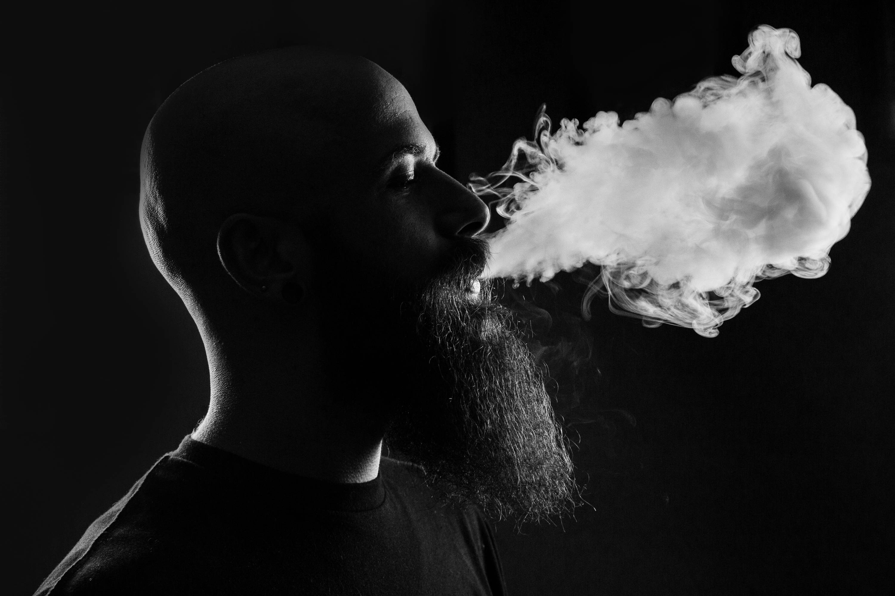 Black and white image of a bald man with a long beard exhaling vape smoke.