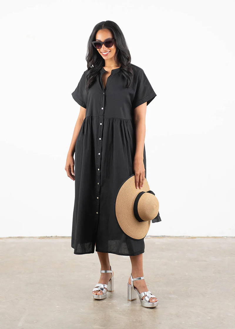 A model wearing an oversized black shortsleeved dress with mother of pearl buttons down the front, silver block heels and holding a wide brimmed hat