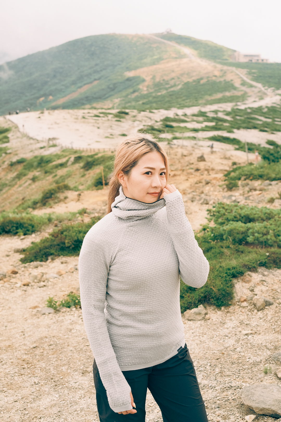 Person standing in nature, wearing a gray sweater, with a green landscape behind.