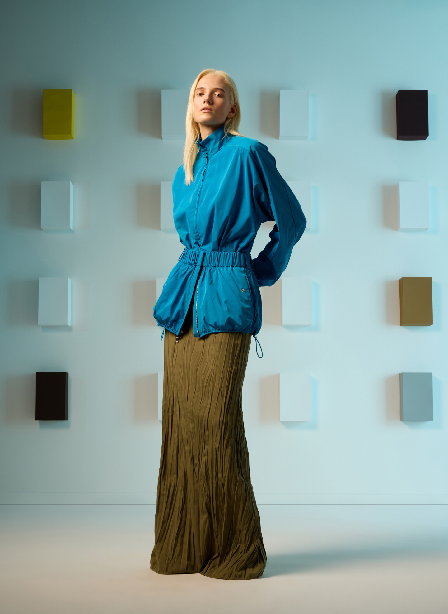Model in blue jacket and green skirt, standing against a colorful background.