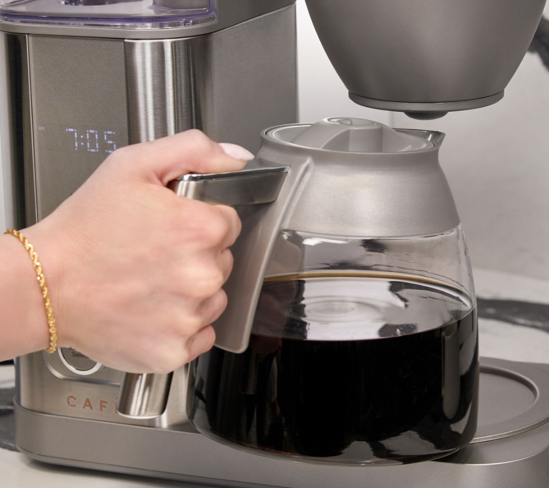 Hand holding a coffee pot, brewing coffee machine in the background.