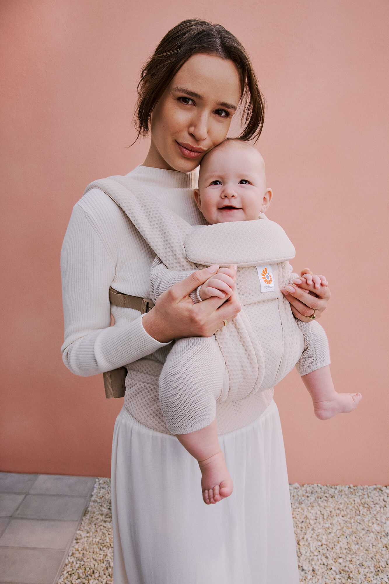 Woman holding a baby in a carrier against a pink background.