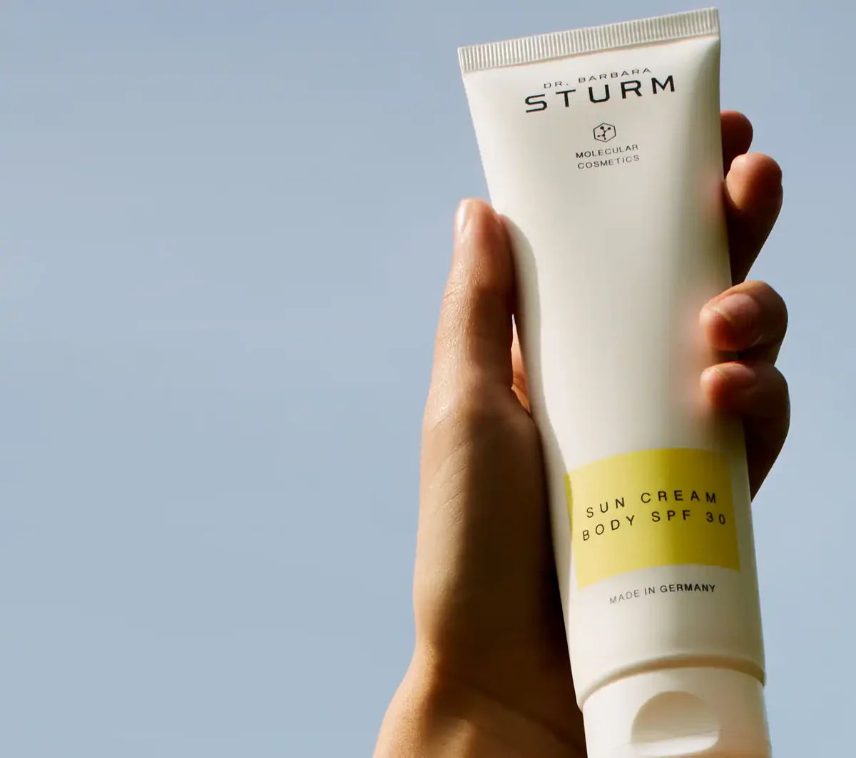 A hand holding a tube of STURM skincare product against a blue sky.
