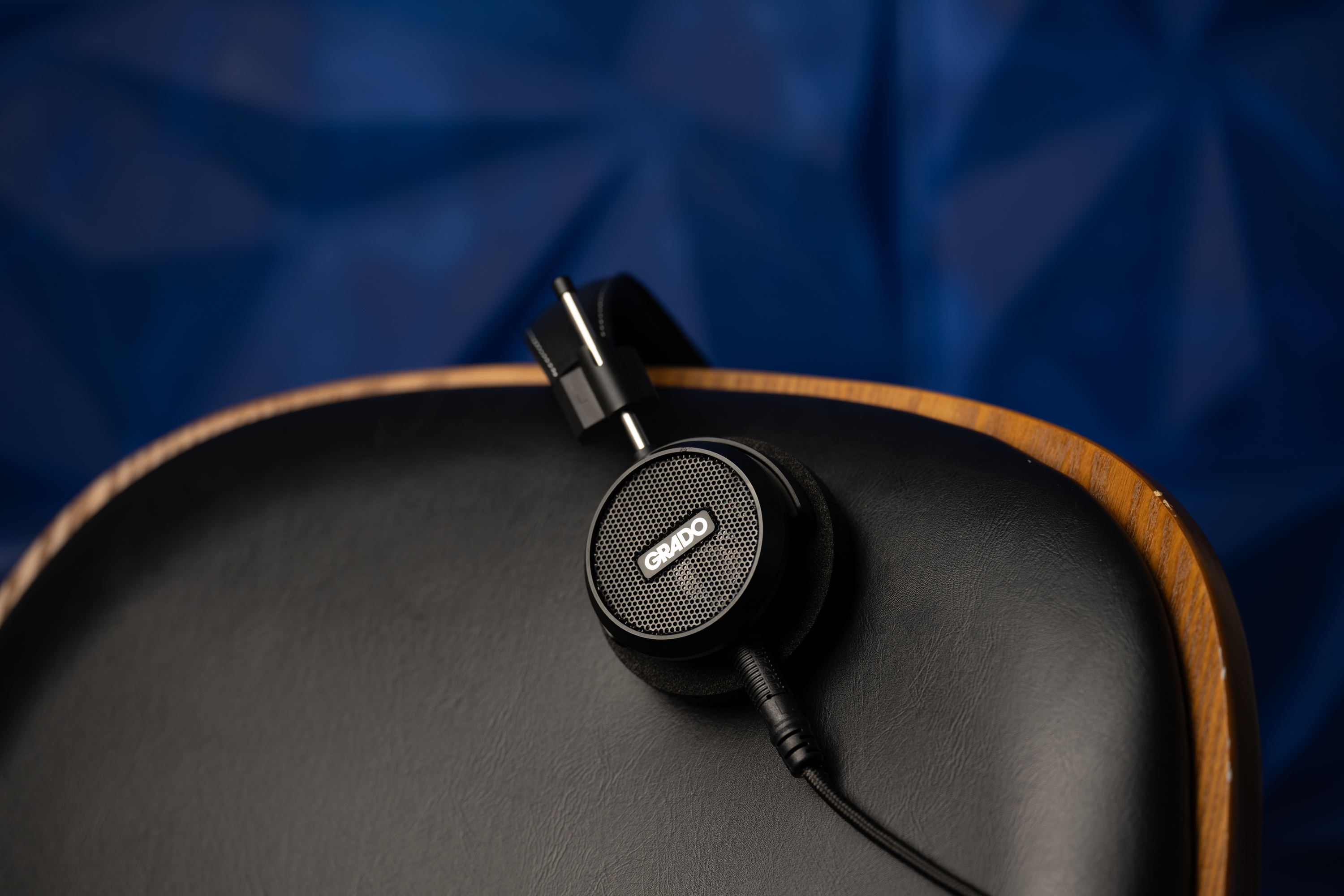 Photo of the Grado S150 Headphones resting on the back of a black leather chair in front of a blue background.