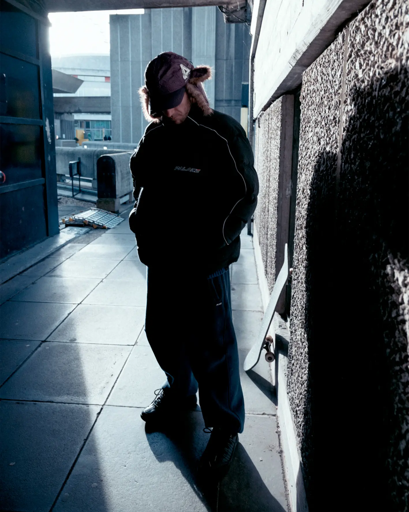 A person in a dark outfit stands in a shadowed urban setting.