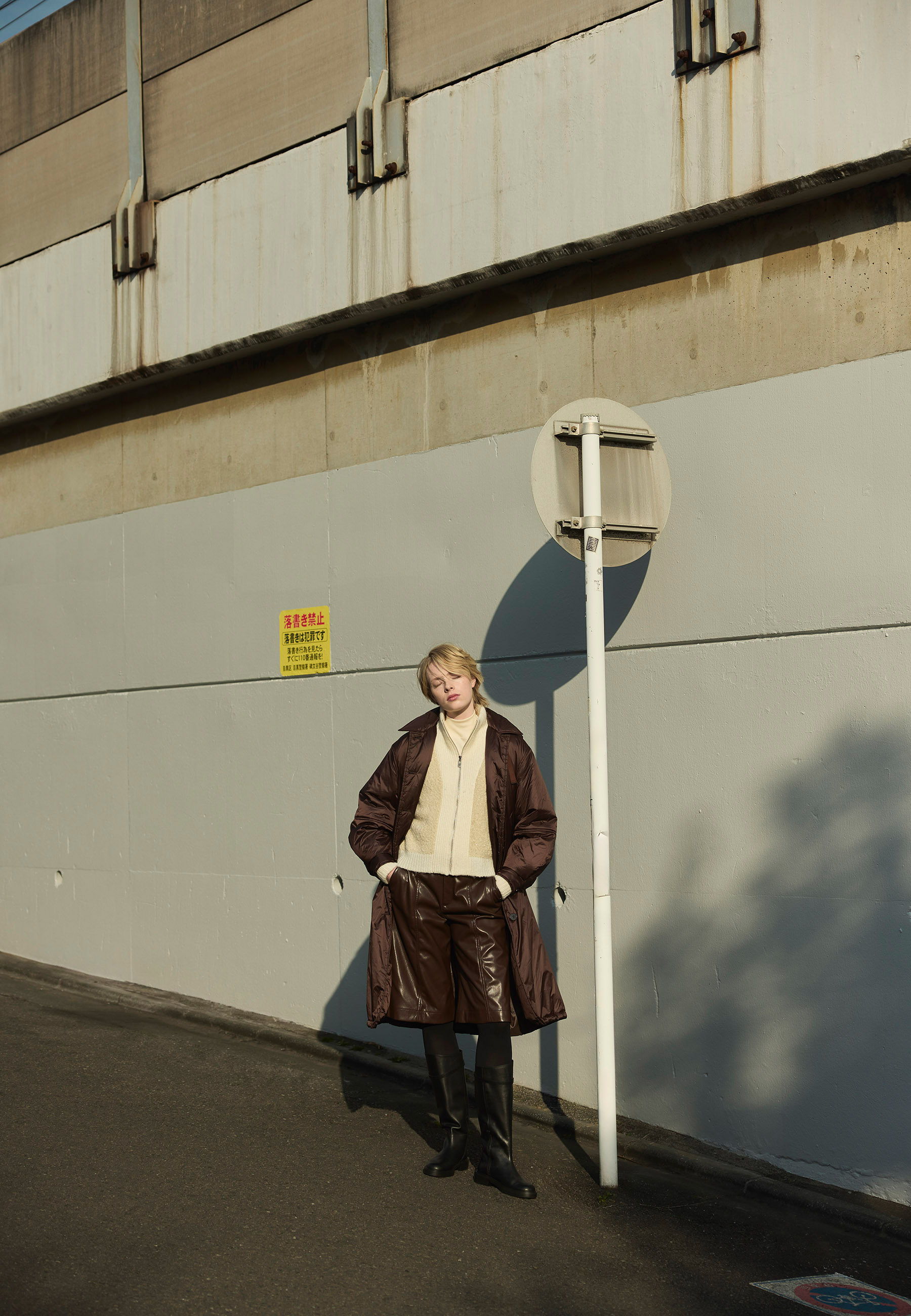 Person in a brown coat stands beside a traffic sign against a gray wall.