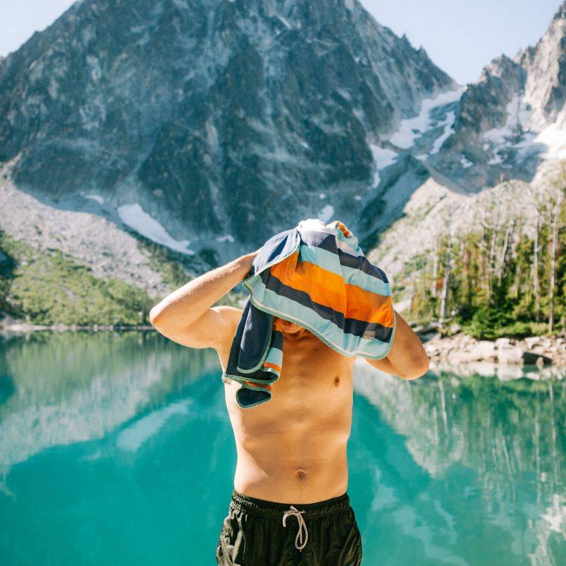 A man holding a towel in front of a lake with mountains in the background.