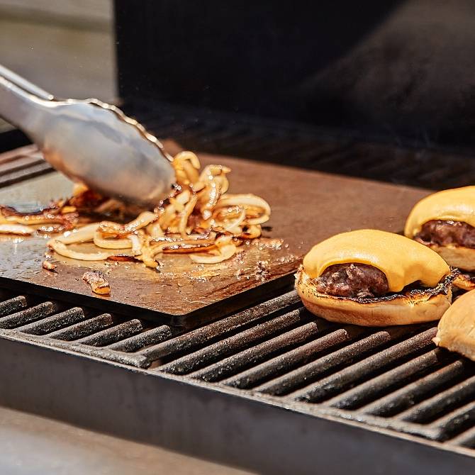 A Misen Oven Steel on an outdoor grill caramelizes thin onion slices while cheeseburgers in buns finish cooking nearby.