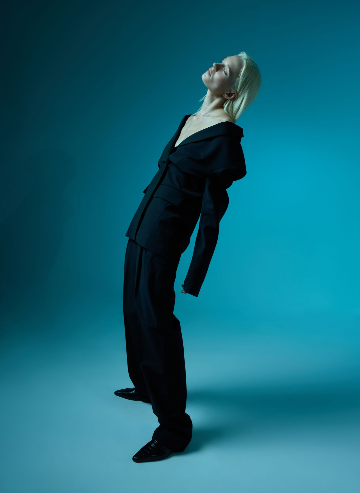Model in a black suit leaning back against a teal background.