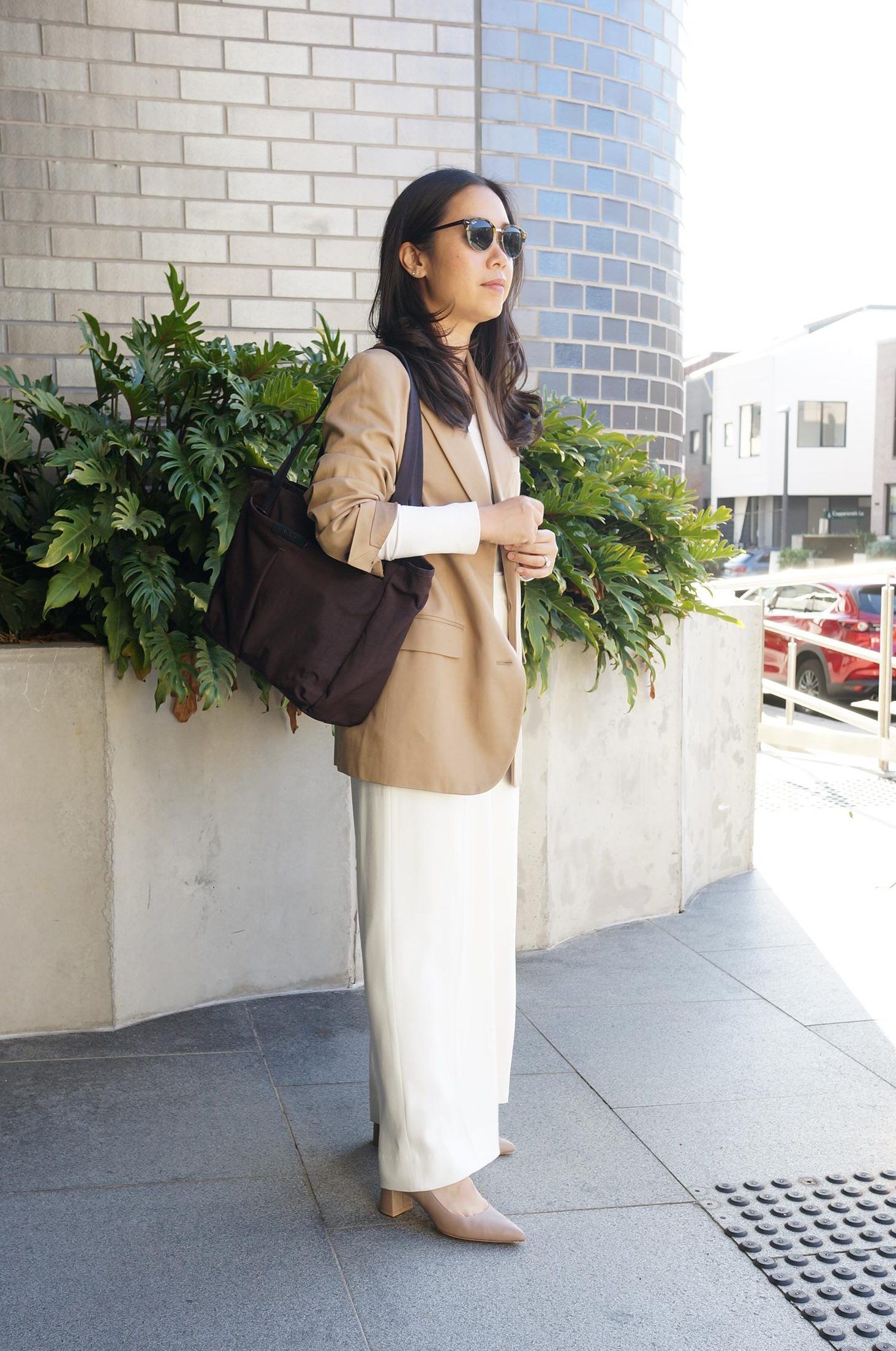 A woman in sunglasses stands outdoors, wearing a blazer and holding a bag.