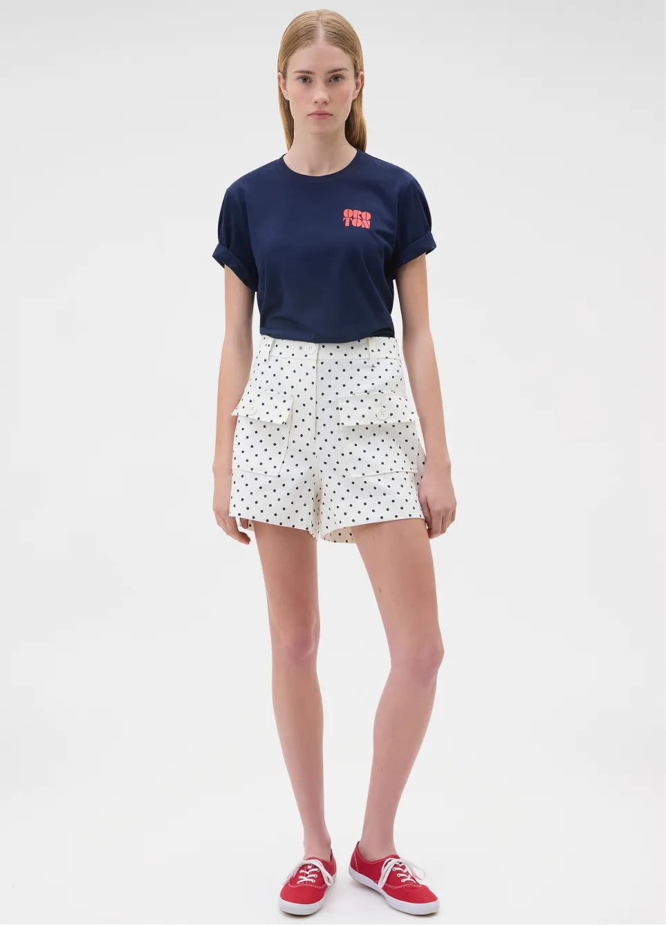 Model wearing a navy t-shirt and polka dot shorts with red shoes.