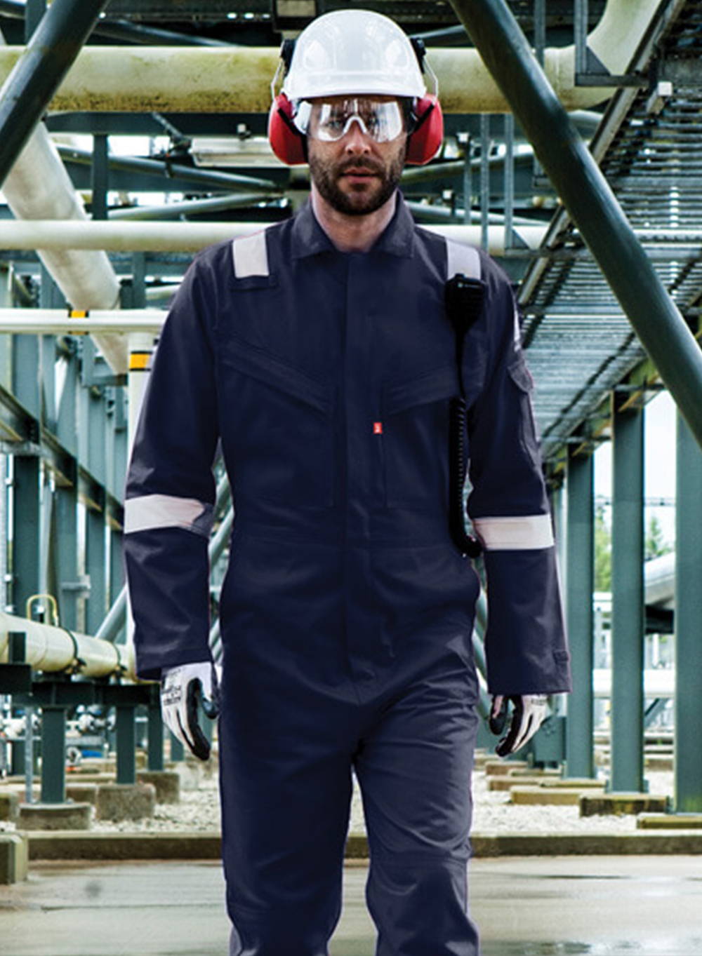 Man walking through tunnel wearing navy coveralls with white hard hat, earmuffs and safety glasses.