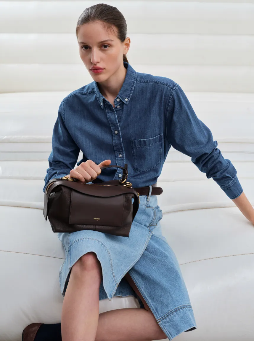 Woman in denim shirt and skirt holding a handbag, seated on a couch.