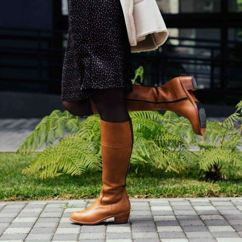 Woman wearing tan leather Condesa wide calf boots with a stacked leather heel, standing on a paved walkway near green ferns.