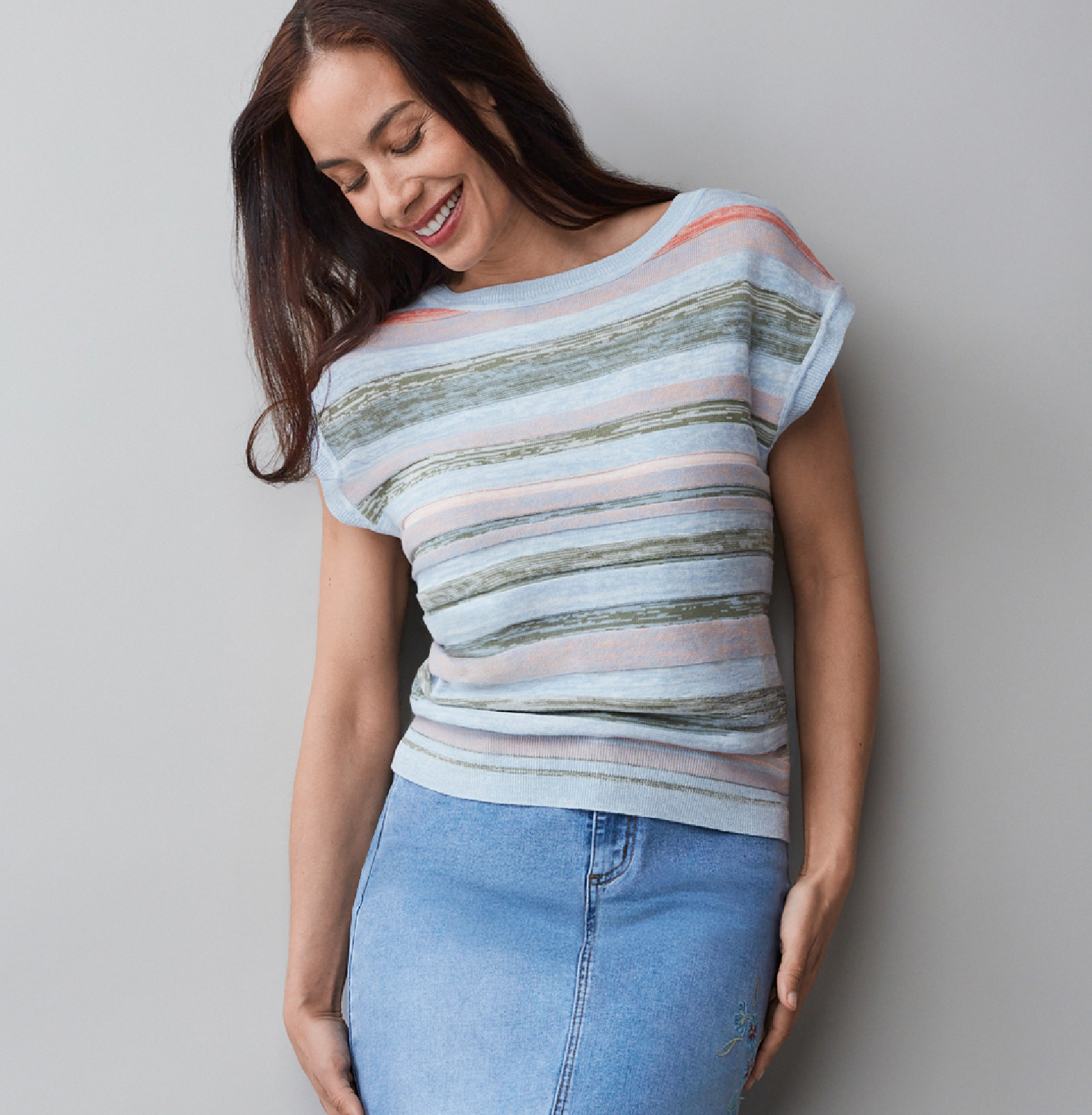 Smiling woman in striped shirt and denim skirt against a gray background.