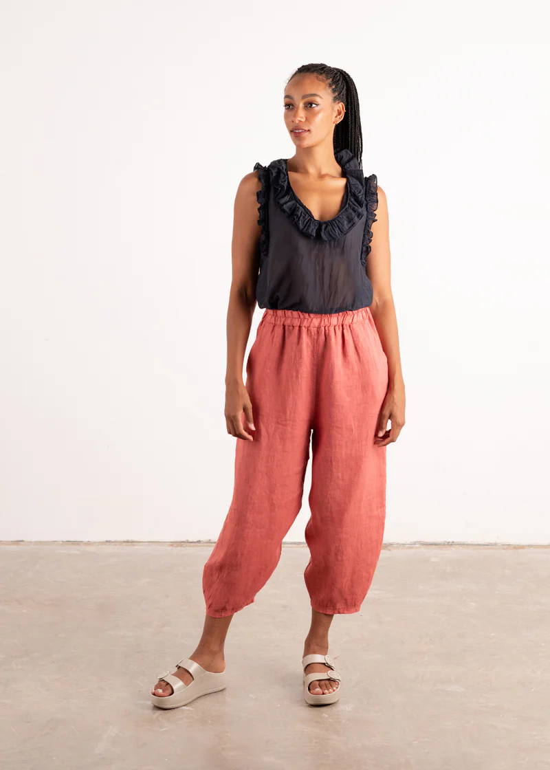 A model wearing a dark blue sleeveless top with frill detail around the neck and arms and dark pink linen barrel leg trousers with off white chunky platform slides 