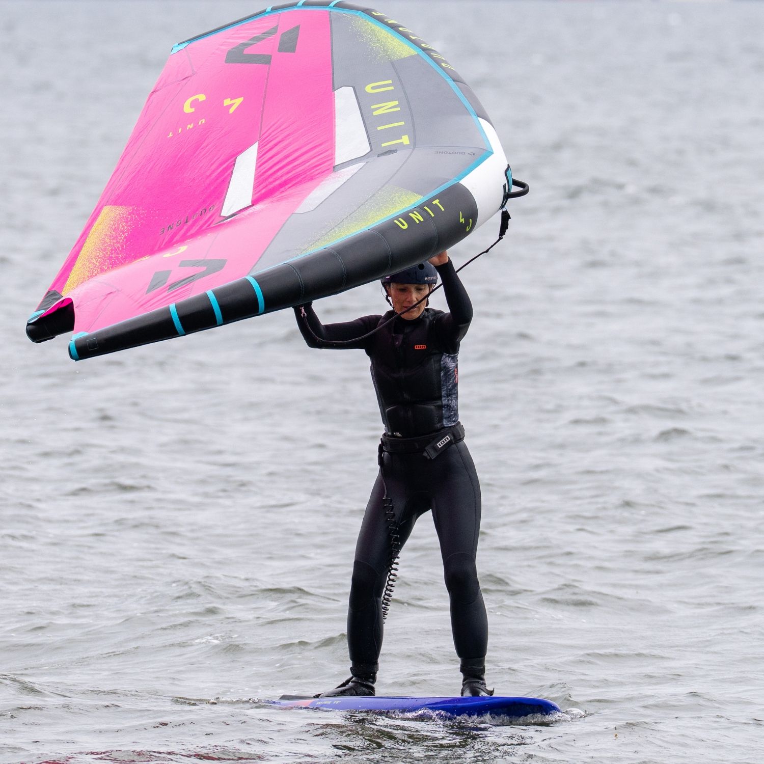 Person in wetsuit holding a colorful sailboard above water.