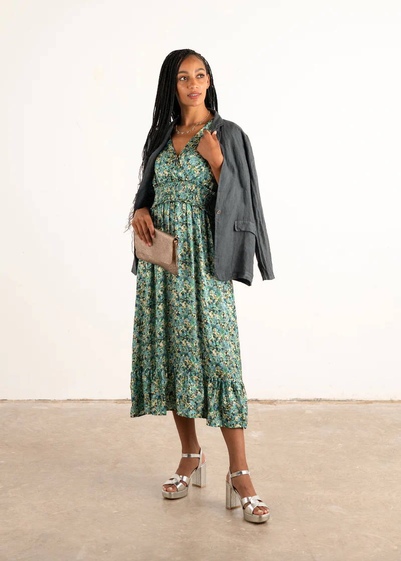 A model wearing a blue satin midi dress with a paisley print with a inky blue linen jacket over her shoulders holding a gold metallic bag and silver chunky heels