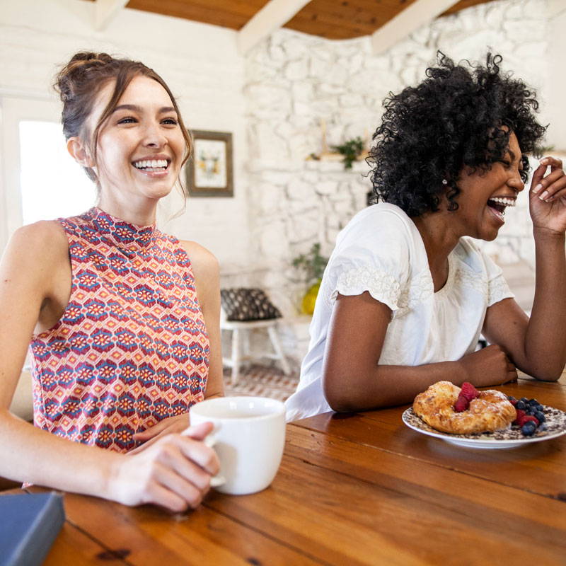 Two female models laughing at breakfast table.
