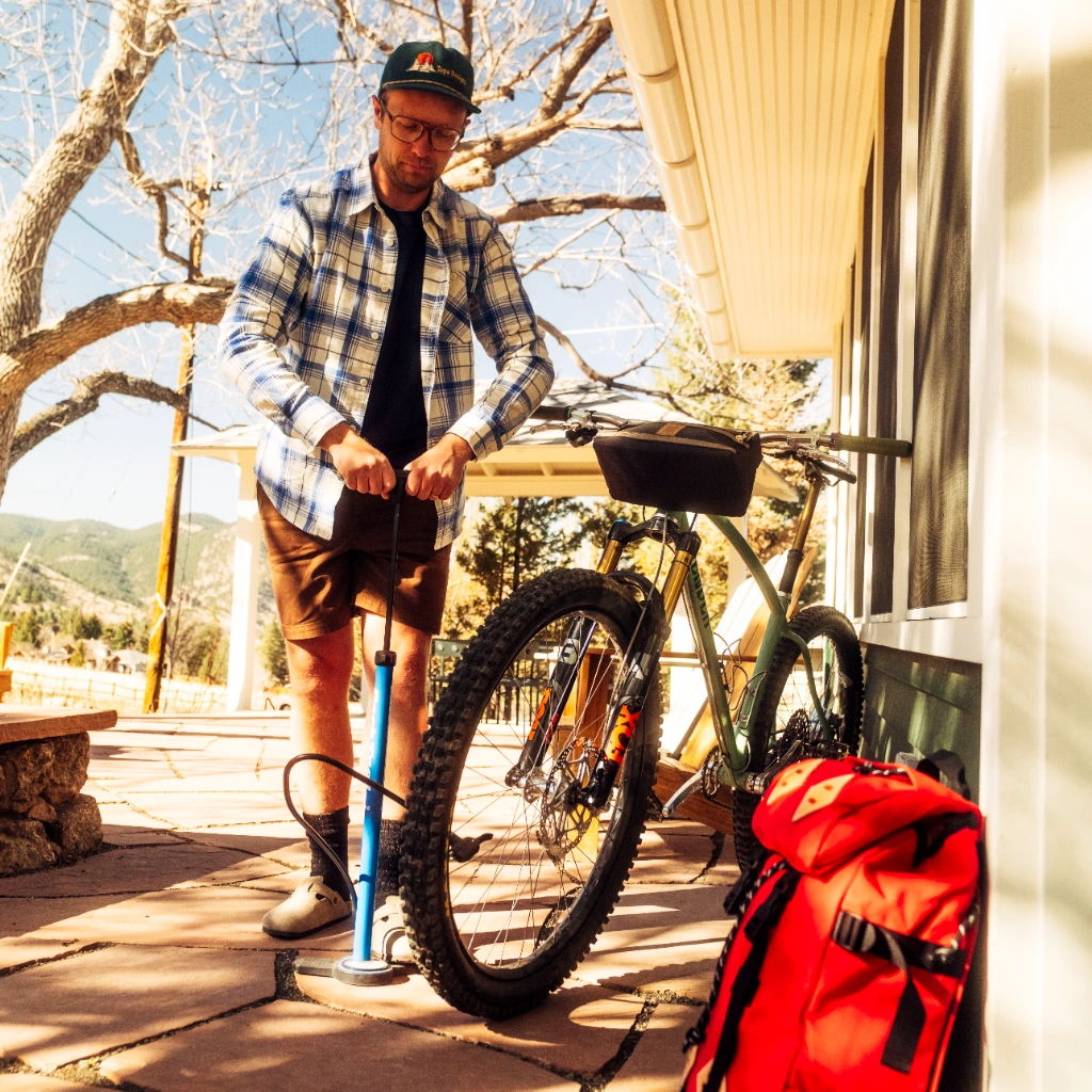 Person inflating a bicycle tire near a house with a red backpack.