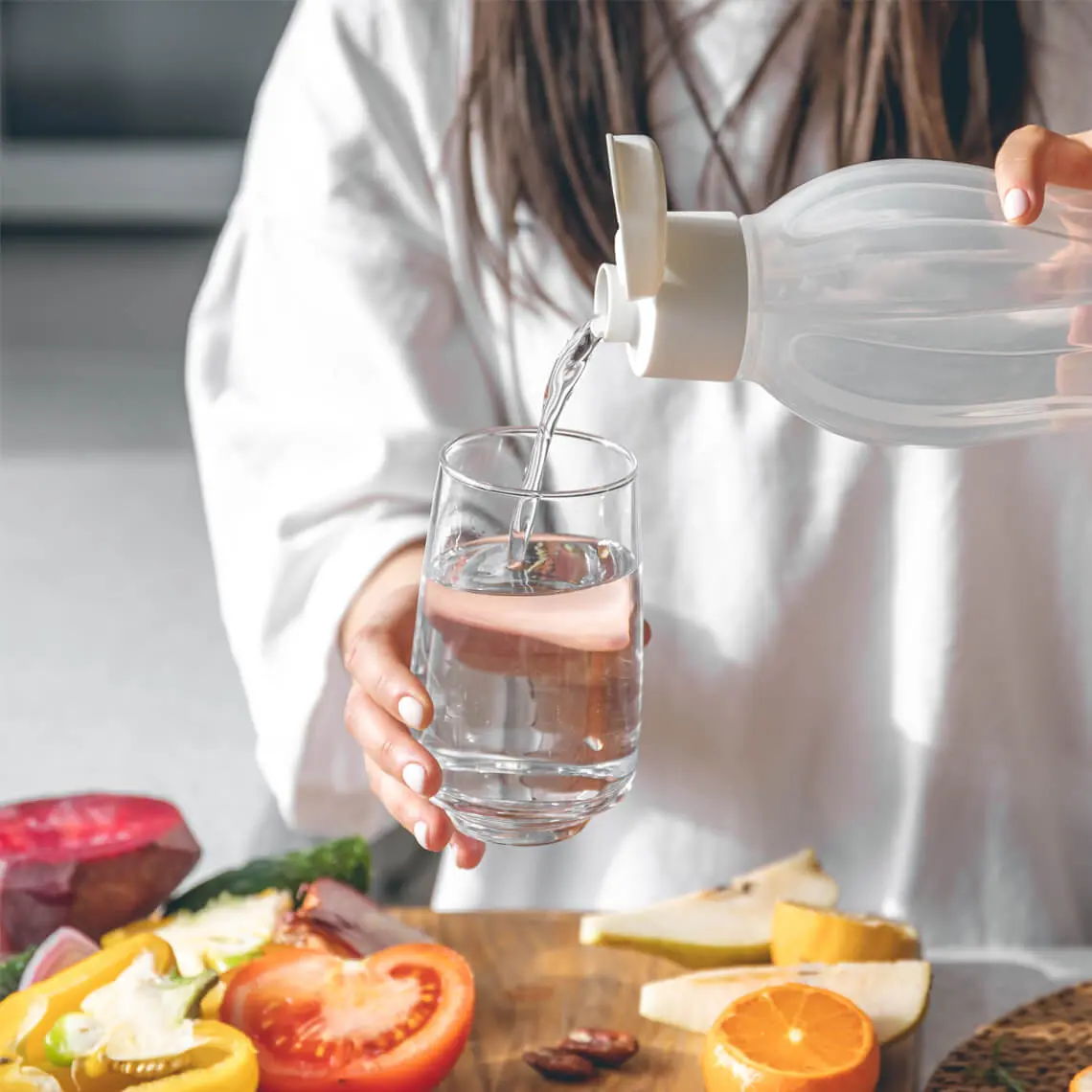 Person pouring water into a glass with fruits on a wooden table.