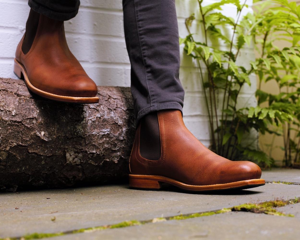 A man standing outside next to a log wearing Adelante brown leather extra wide boots with elastic side panels.