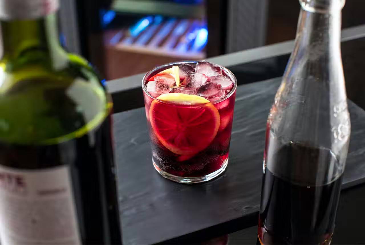 Kalimotxo in a tumbler glass with a slice of lemon. Bottles of wine and soda are also displayed.
