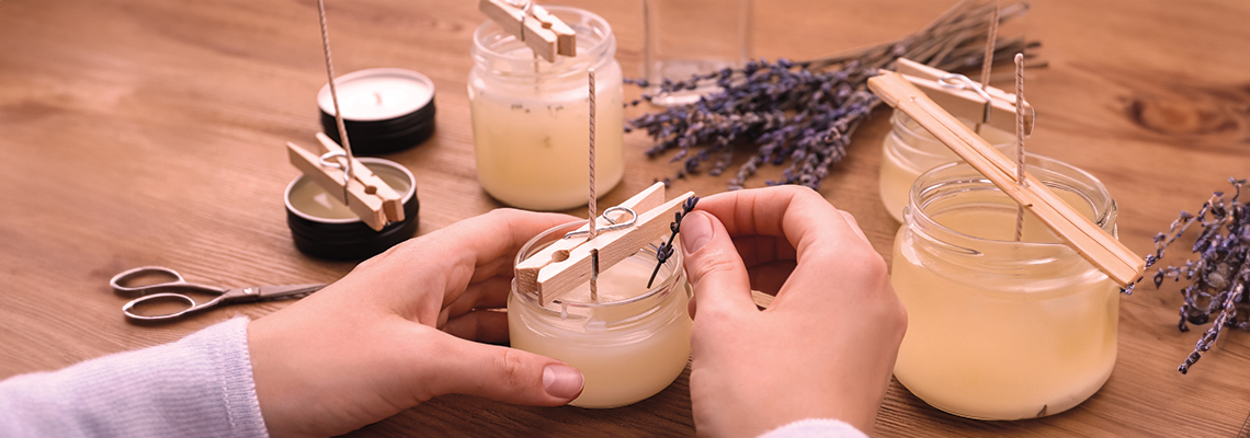 Person making candles with jars, clothespins, and lavender on a wooden table.