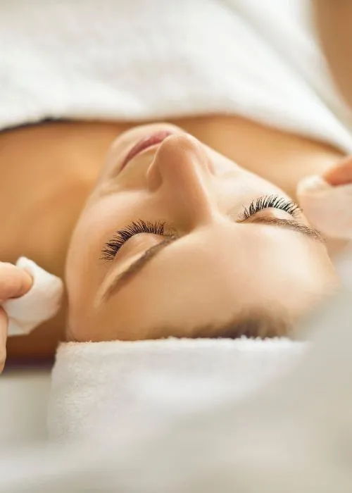 A woman receiving a facial treatment with cotton pads on her eyes.