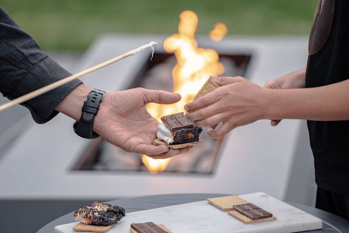 Hands making s’mores beside a modern gas fire pit, with graham crackers and chocolate on a tray