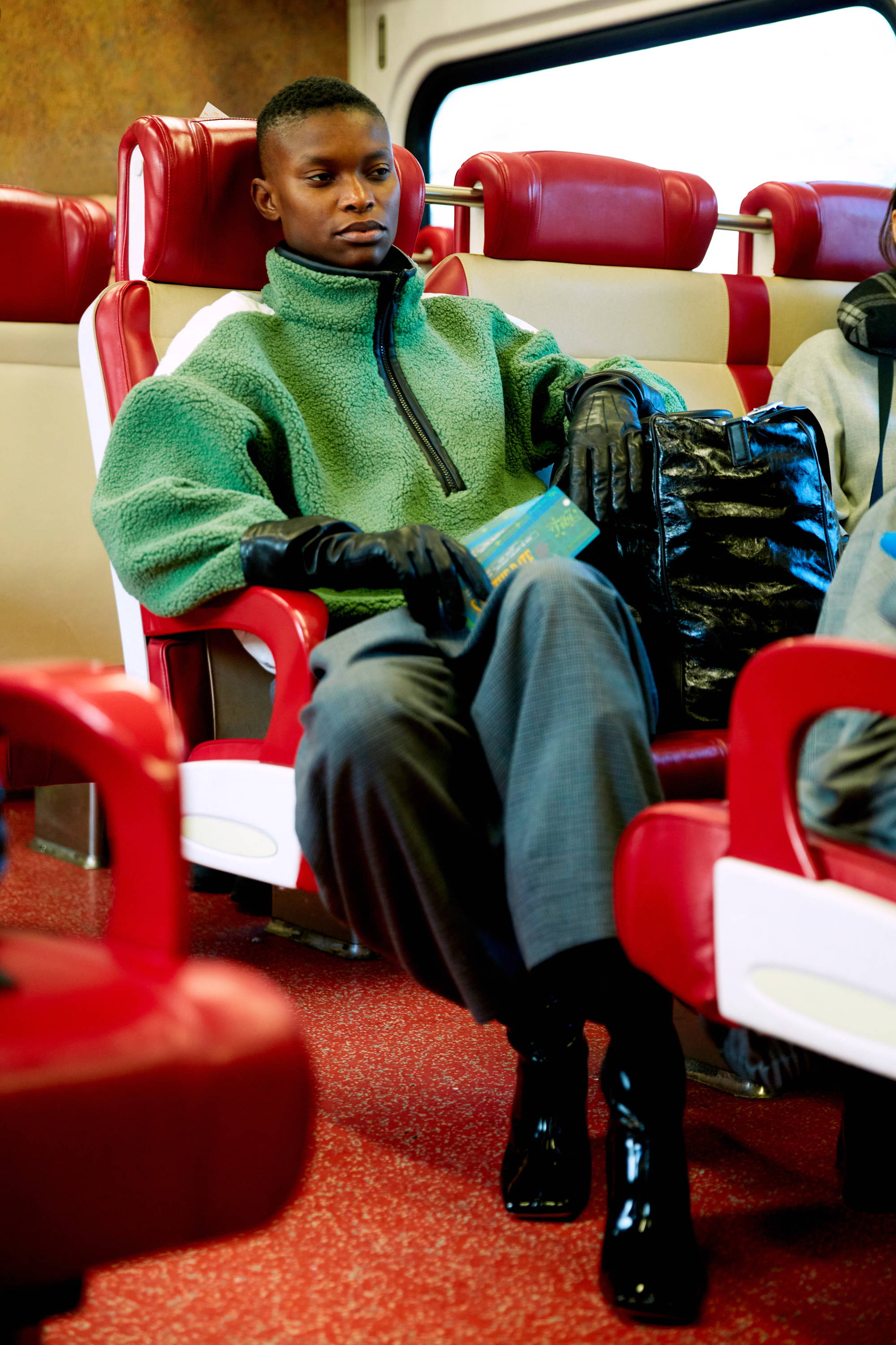 A person in a green sweater sits on a train seat, wearing gloves.
