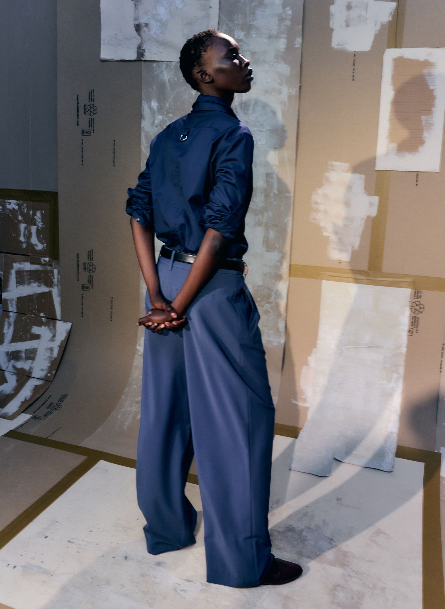 Person in dark outfit standing with hands behind back, against abstract background.