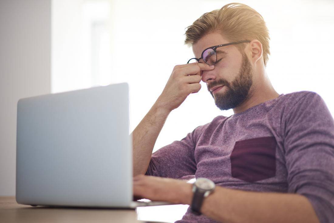 Man sitting at a computer, rubbing his eyes showing symptoms of eyestrain wearing a purple shirt with a black watch