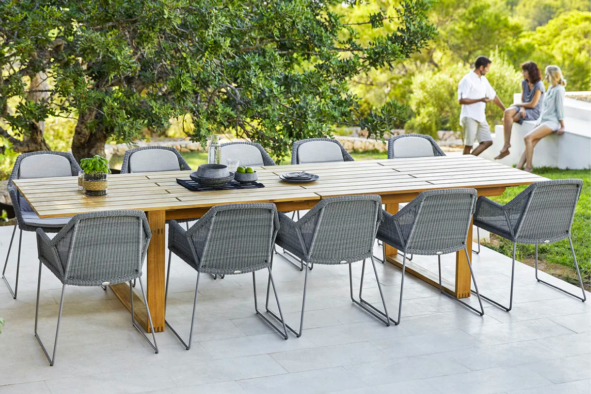 An expansive teak outdoor table with woven grey chairs and people chatting in the background.
