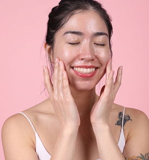 Young woman smiling and applying skincare on her face against a pink background.