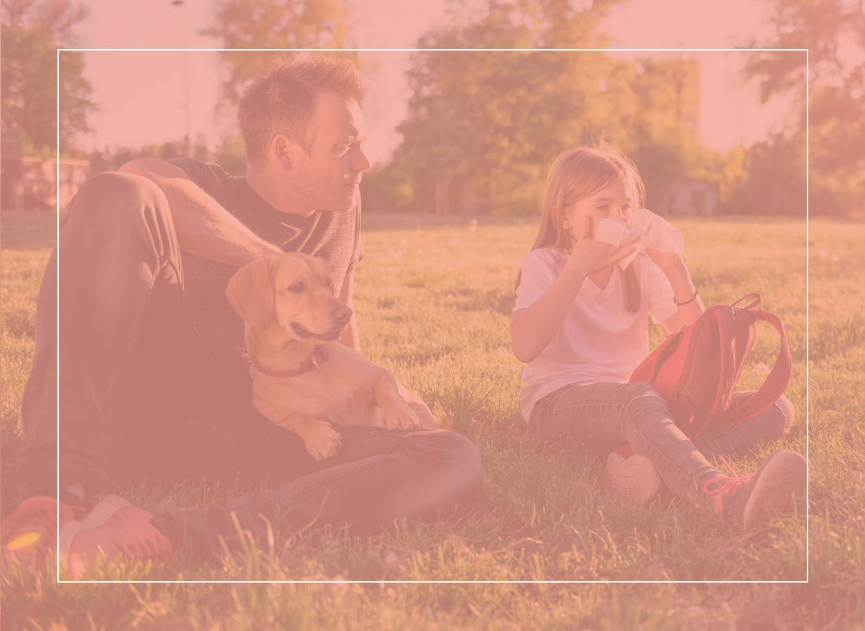 Dívka s alergií na pyl smrká. Sedí během letního dne v trávě se svým otcem a psem.