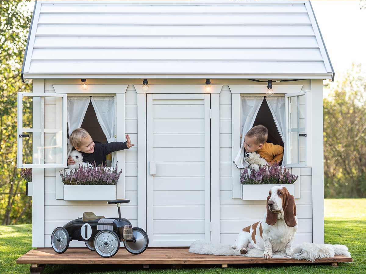 Wooden Playhouse in white color with two boys at the windows and a car and a big dog in front by WholeWoodPlayhouses