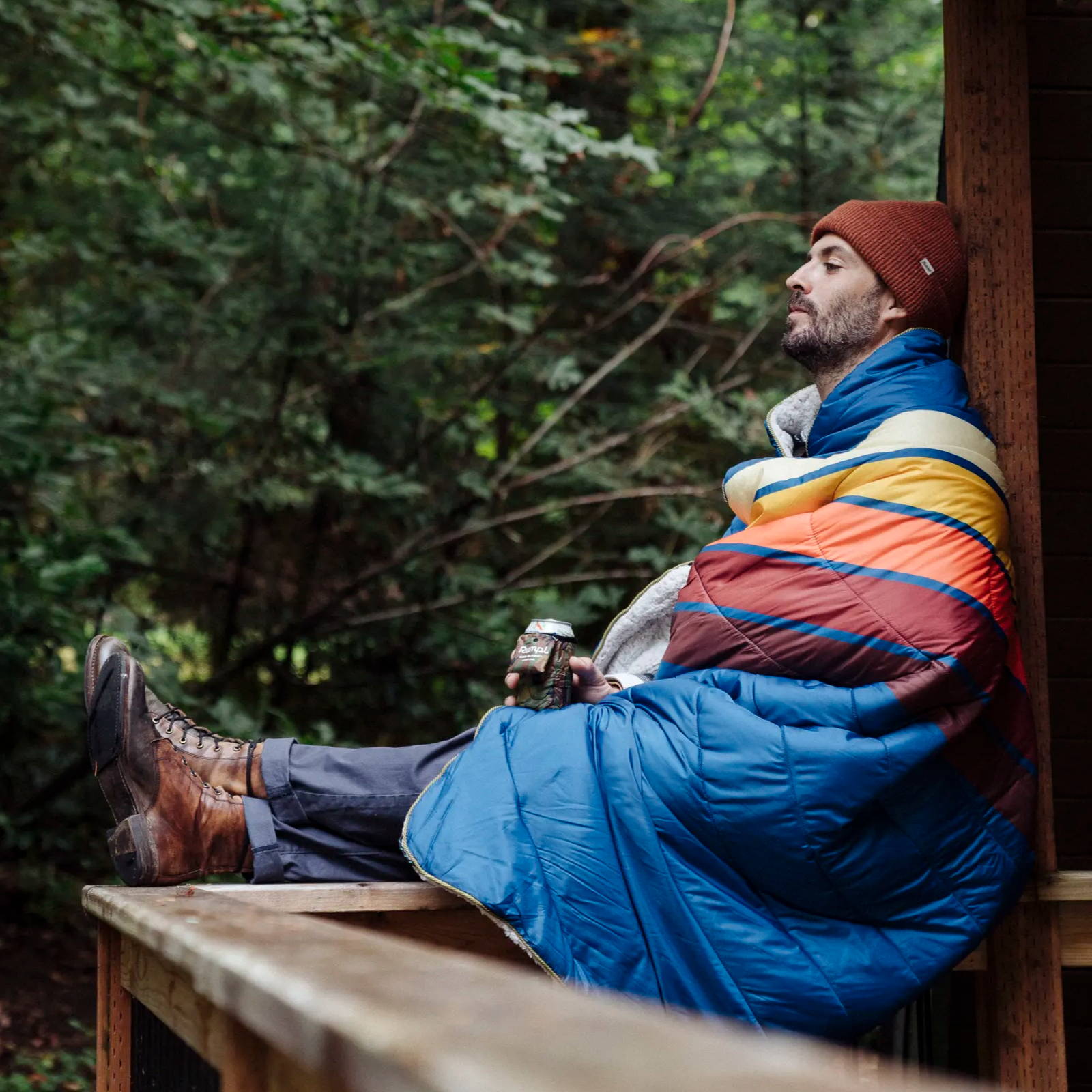 Man sitting on the porch of a cabin wrapped in a sherpa fleece Rumpl blanket drinking a beer