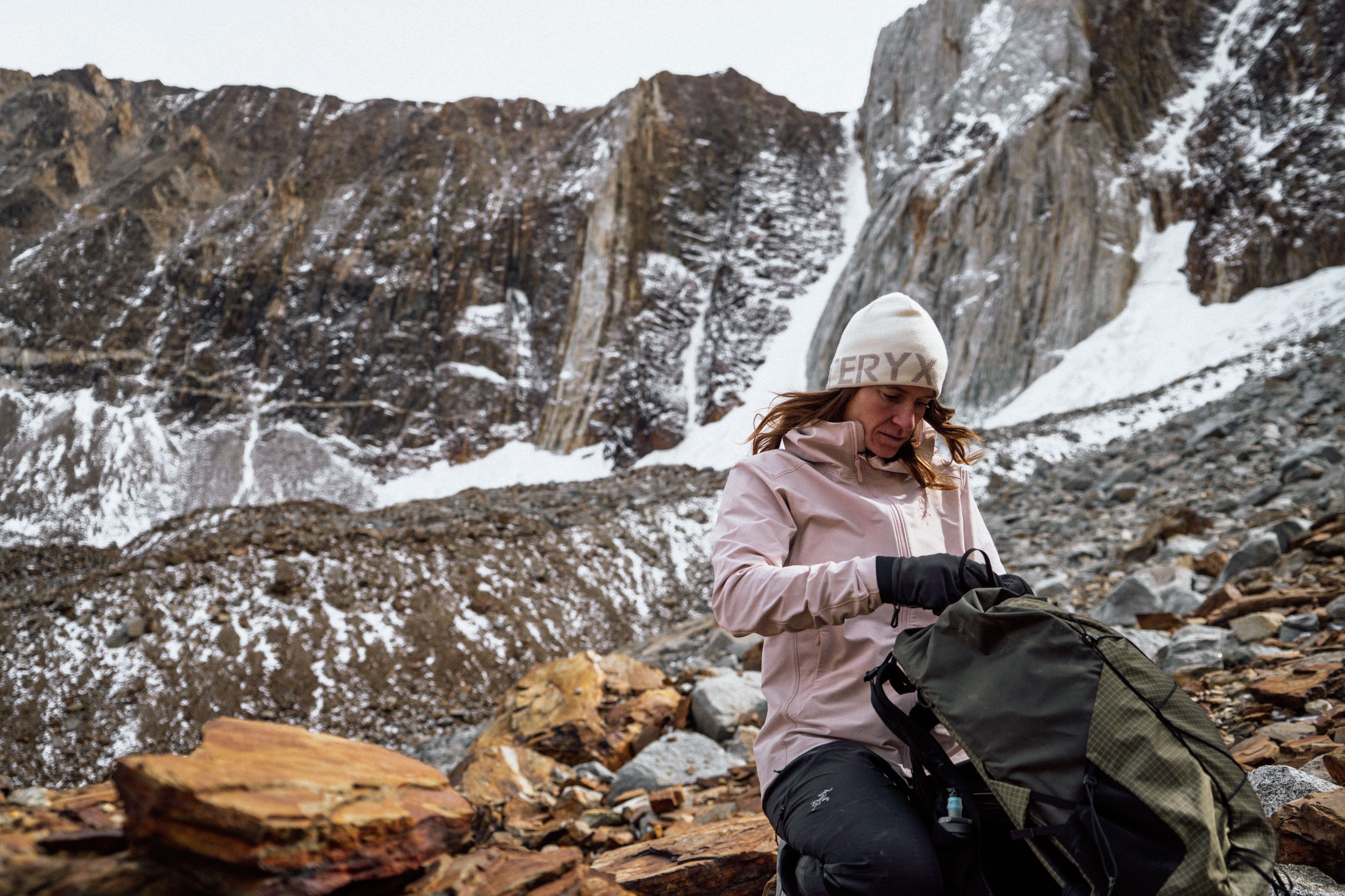 Person in winter clothing adjusting a backpack on a rocky, snowy landscape.