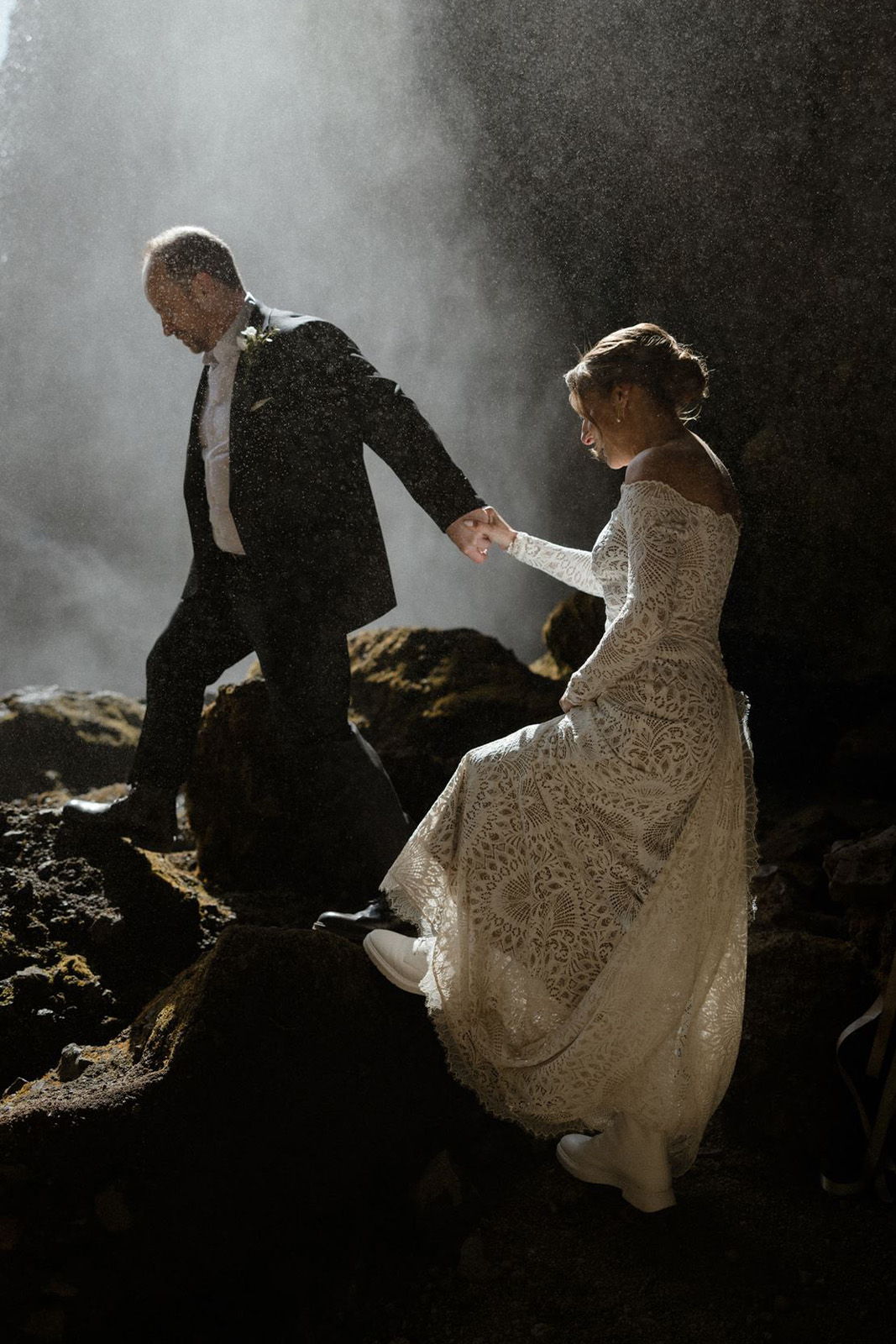 Couple in wedding attire walking hand in hand on rocky terrain.