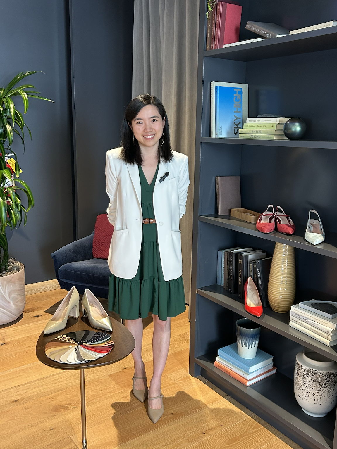 Woman in a green dress and white blazer stands beside a bookshelf.