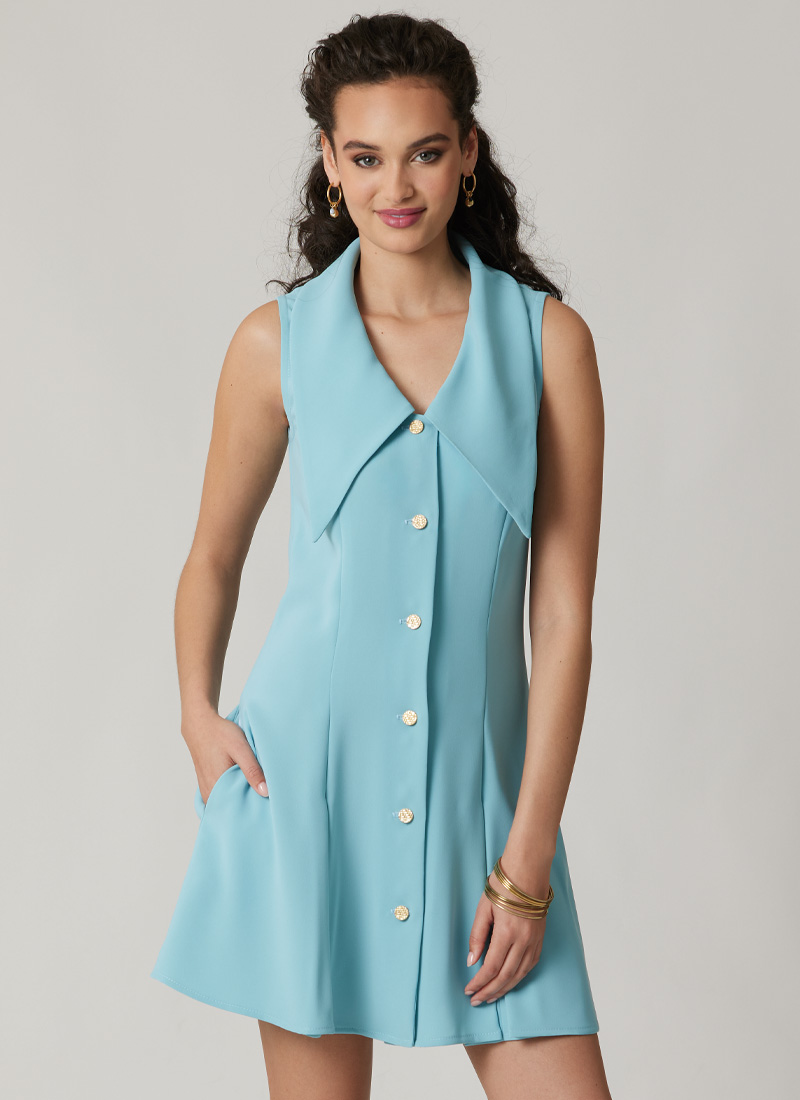 Woman in a sleeveless blue dress with buttons, standing and smiling.