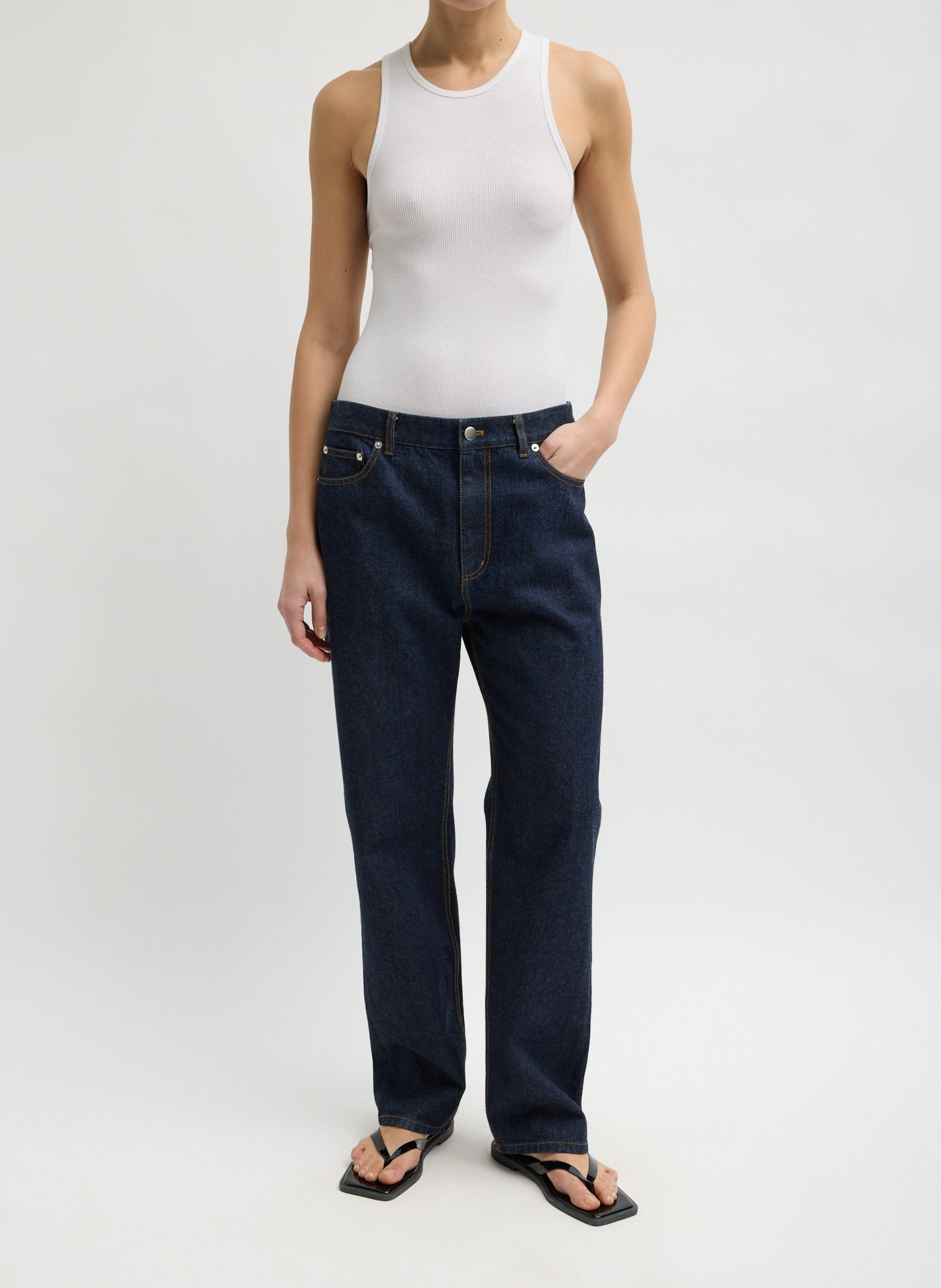Person wearing a white tank top and dark jeans, standing against a neutral background.