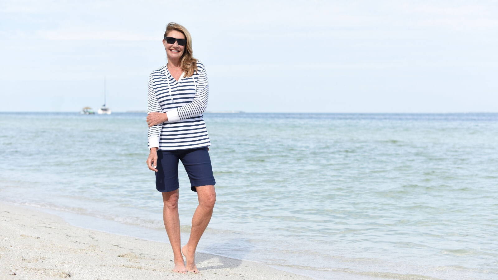 A blonde woman in a striped top, shorts, and sunglasses smiles as she wades in the water at the beach 