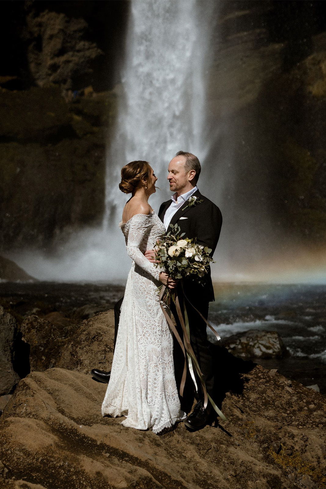 A couple in formal attire embracing near a waterfall.