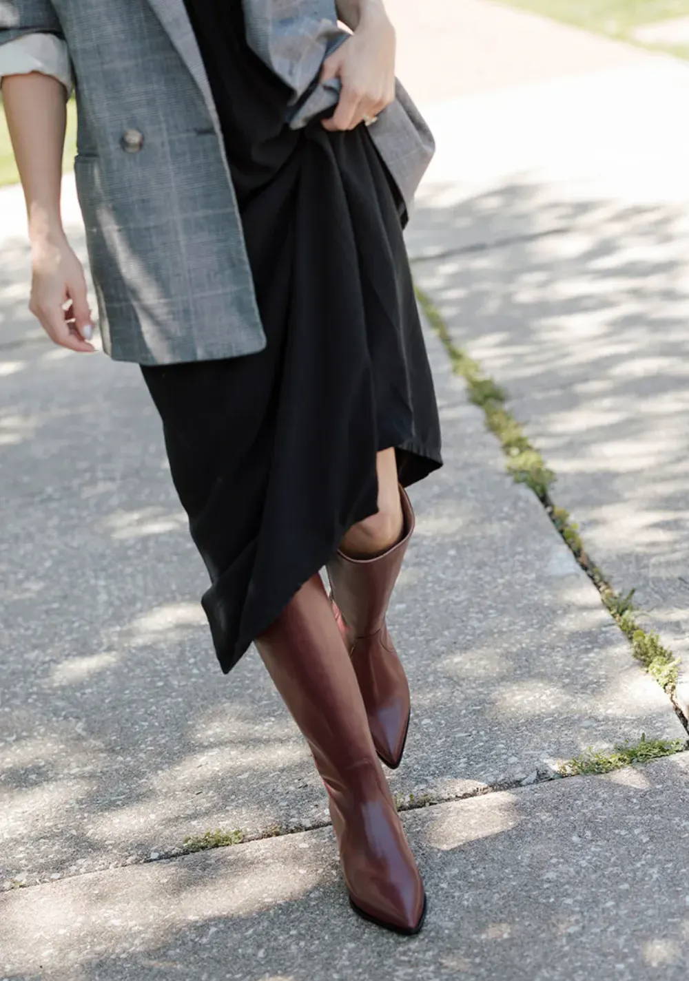 A woman wearing a blazer and dress with brown boots on a sidewalk.