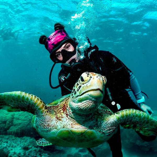 Sally Higgs in scuba gear, wearing a face shield as a headband, posing with a sea turtle.