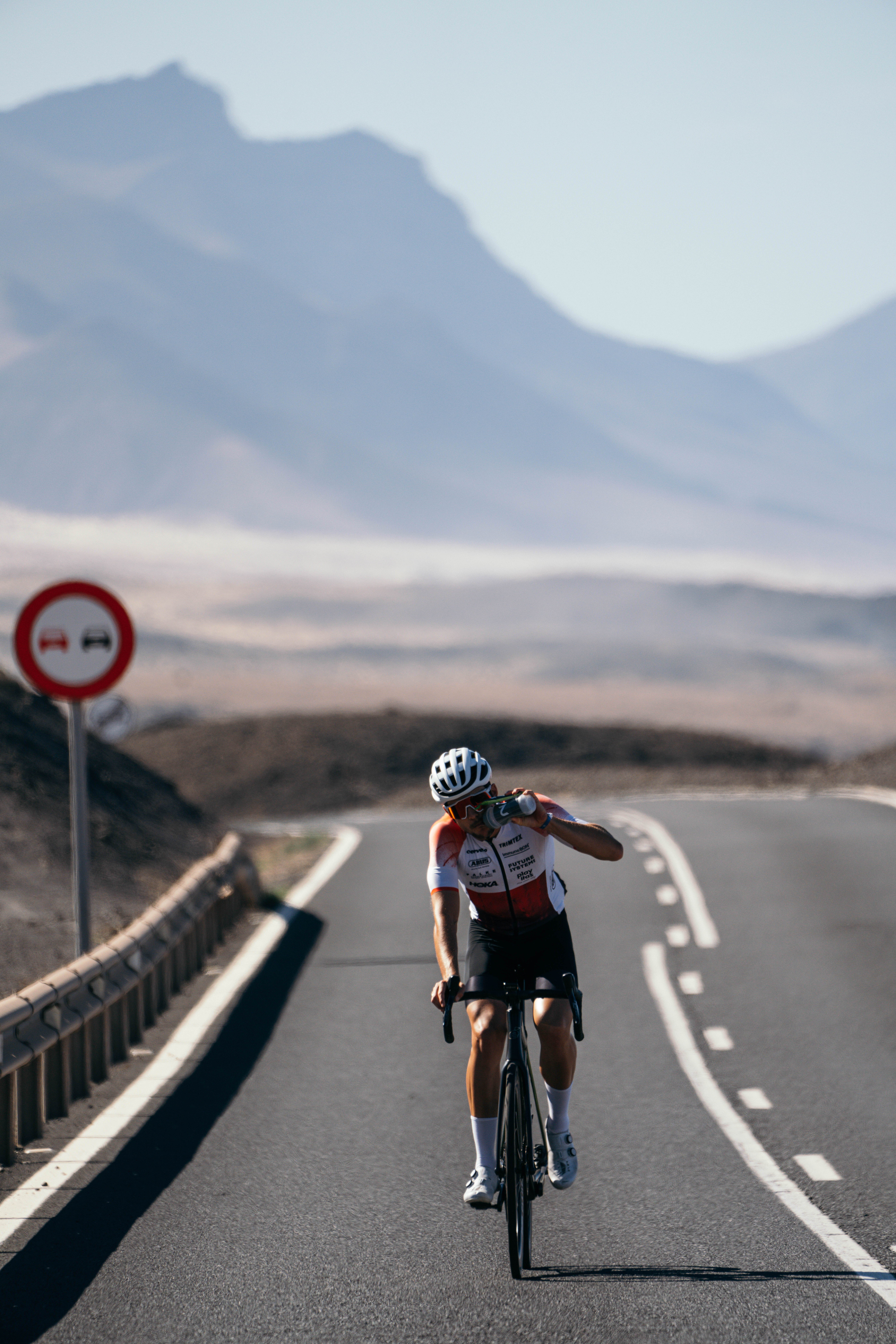 Triathlet Jan Stratmann fährt auf einer abgelegenen Bergstraße und trinkt während der Fahrt aus seiner Flasche von Neversecond.