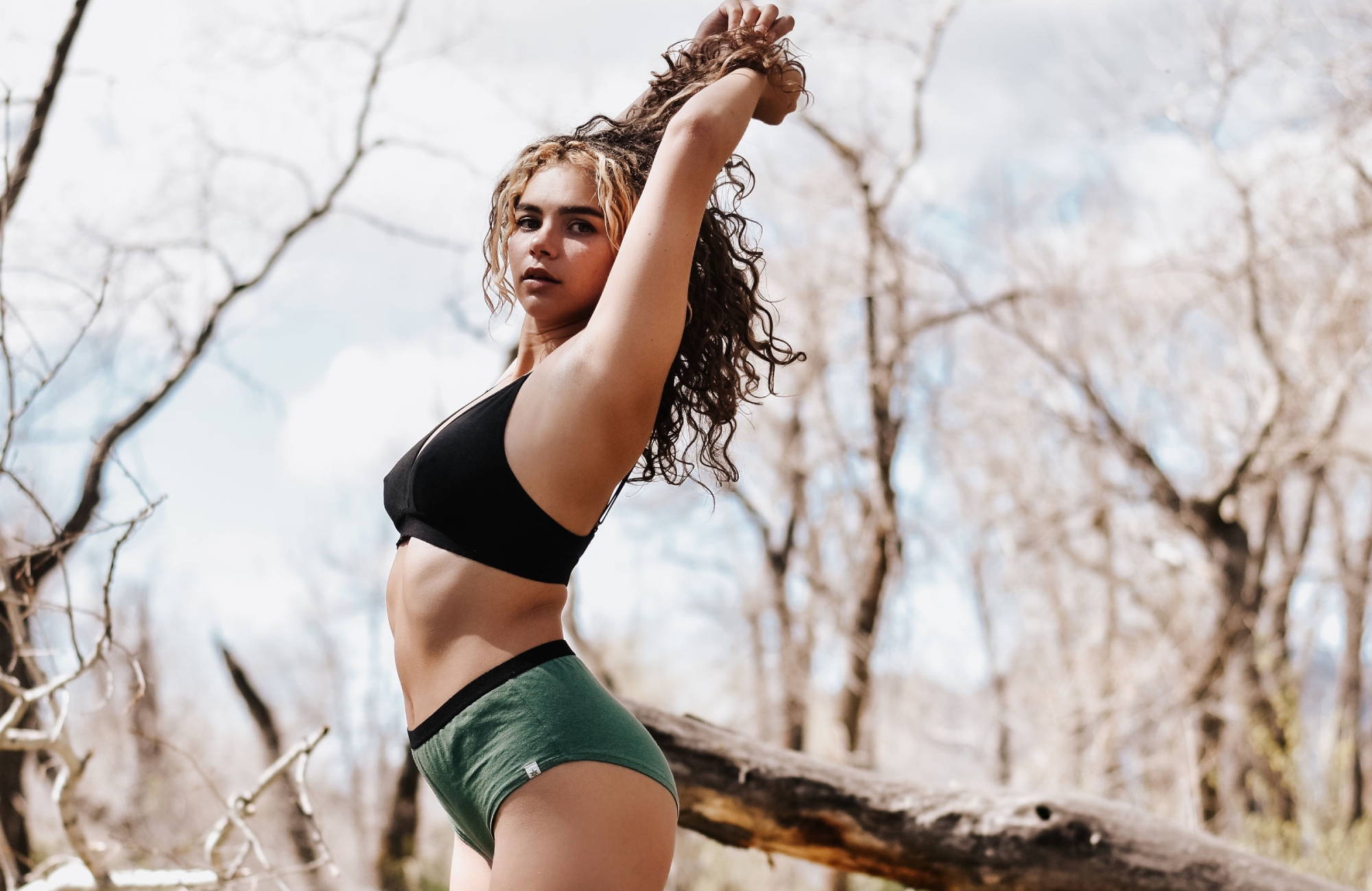 curly haired woman holds her hair up in the woods wearing a black bra and green underwear